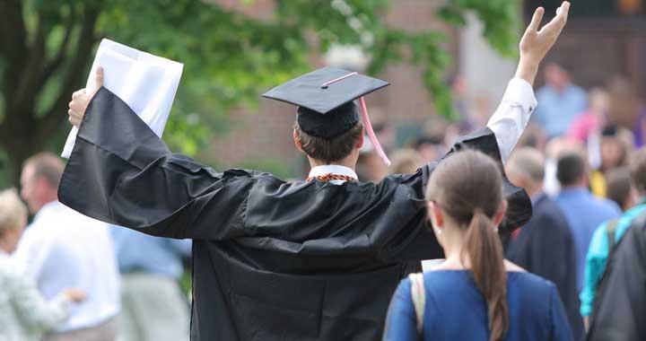 man in graduation cap holding hands up in air outside around other people